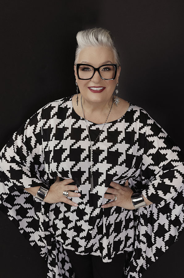 Pegine stands confidently with hands on hips. She wears a striking black and white houndstooth-patterned top and black-rimmed glasses.